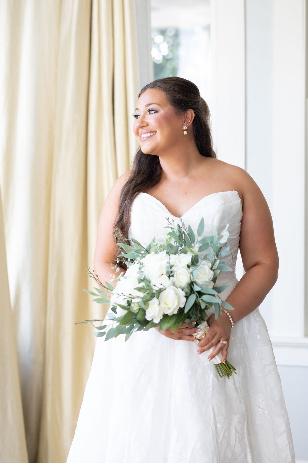 Radiant bride in strapless lace gown holding a white rose and eucalyptus bouquet by a window.