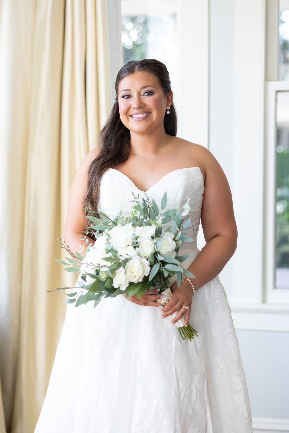 Happy bride in strapless white lace gown smiling while holding a white rose bridal bouquet.