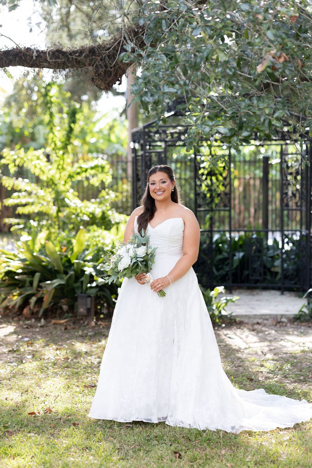 Bride in strapless lace A-line wedding gown holding white floral bouquet, posing outdoors near iron gate and lush greenery.