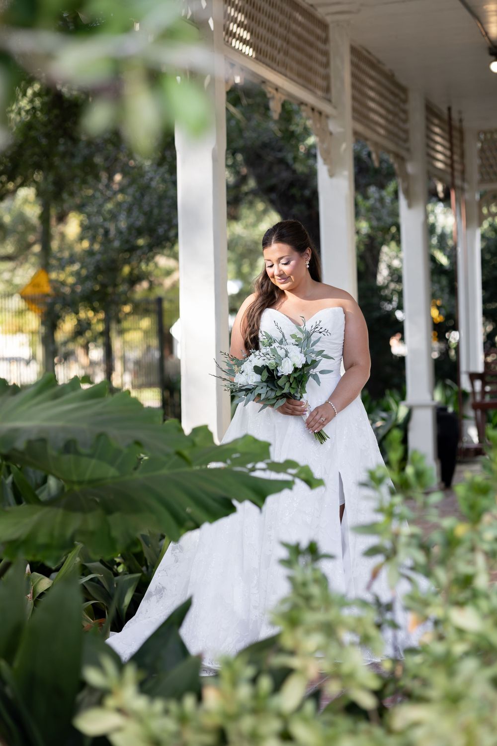 Elegant bride in strapless lace wedding gown holding white bouquet on sunlit colonial porch.