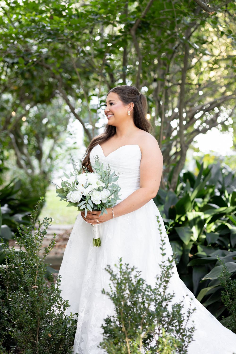 Smiling bride in strapless lace wedding gown holding white bouquet with greenery in lush garden setting.