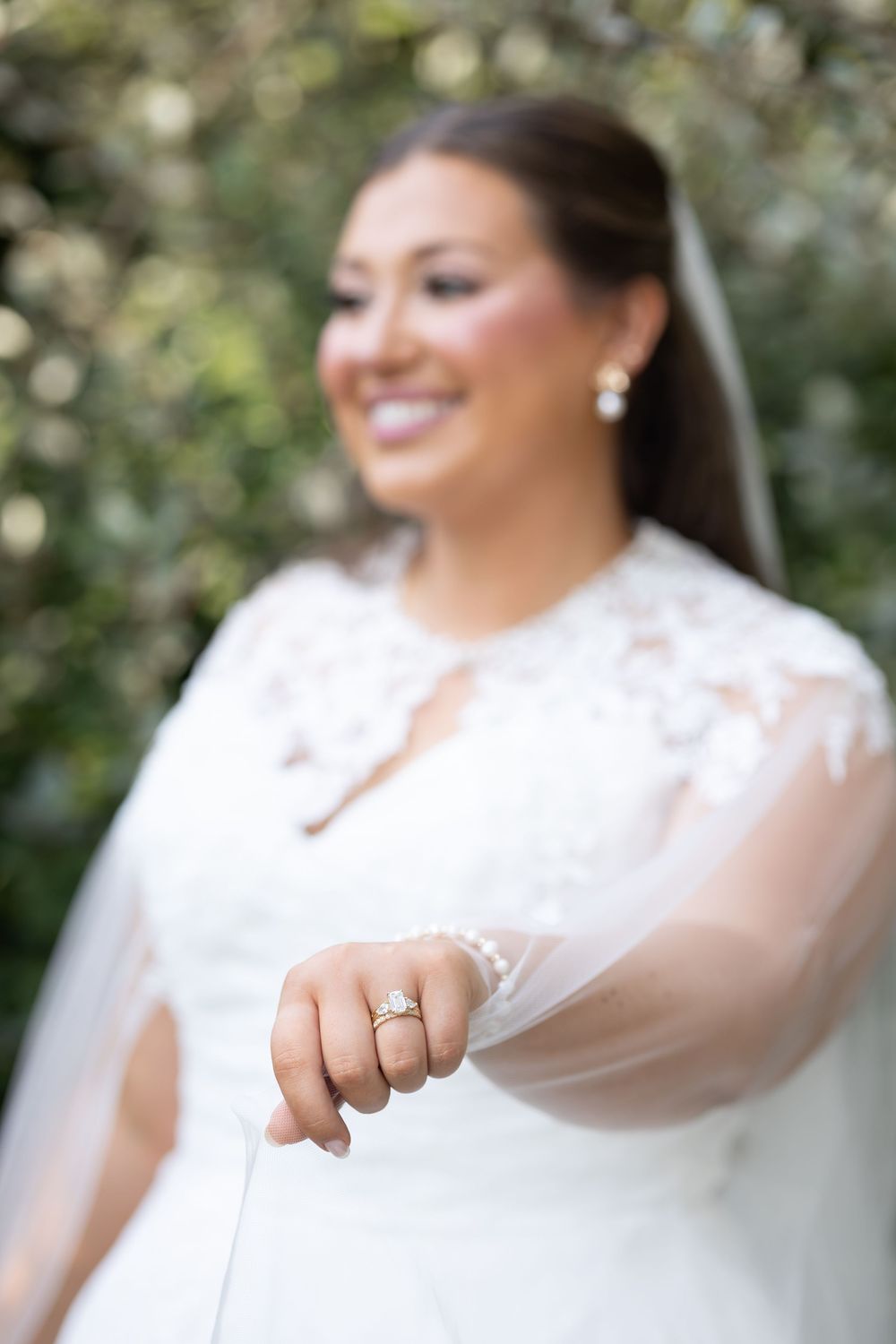 Close-up of smiling bride showing off sparkling diamond engagement ring with veil and lace gown in garden setting.