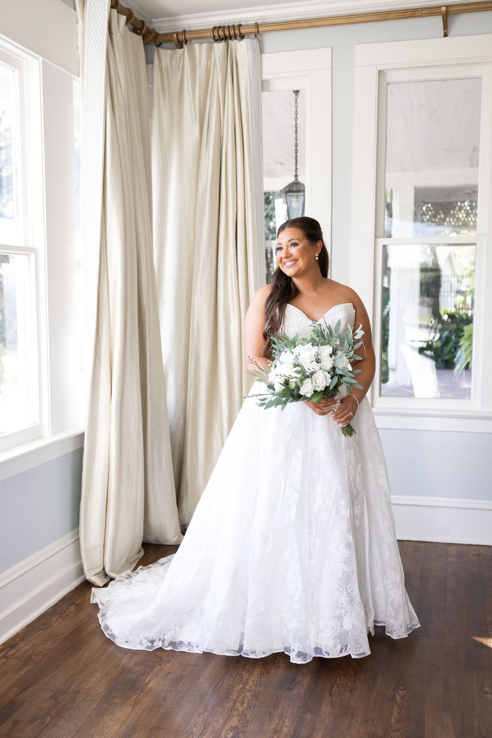 Smiling bride in strapless white ball gown holding white and greenery bouquet standing by elegant curtained windows.
