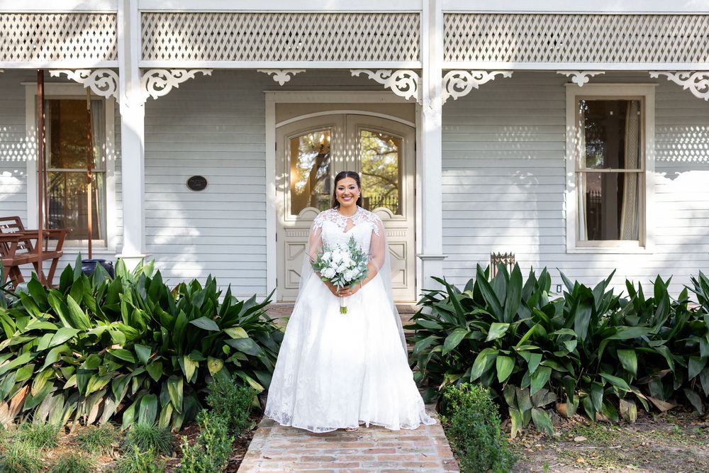 Bride in white wedding gown holding bouquet stands on brick path before charming white Victorian porch with lush greenery.