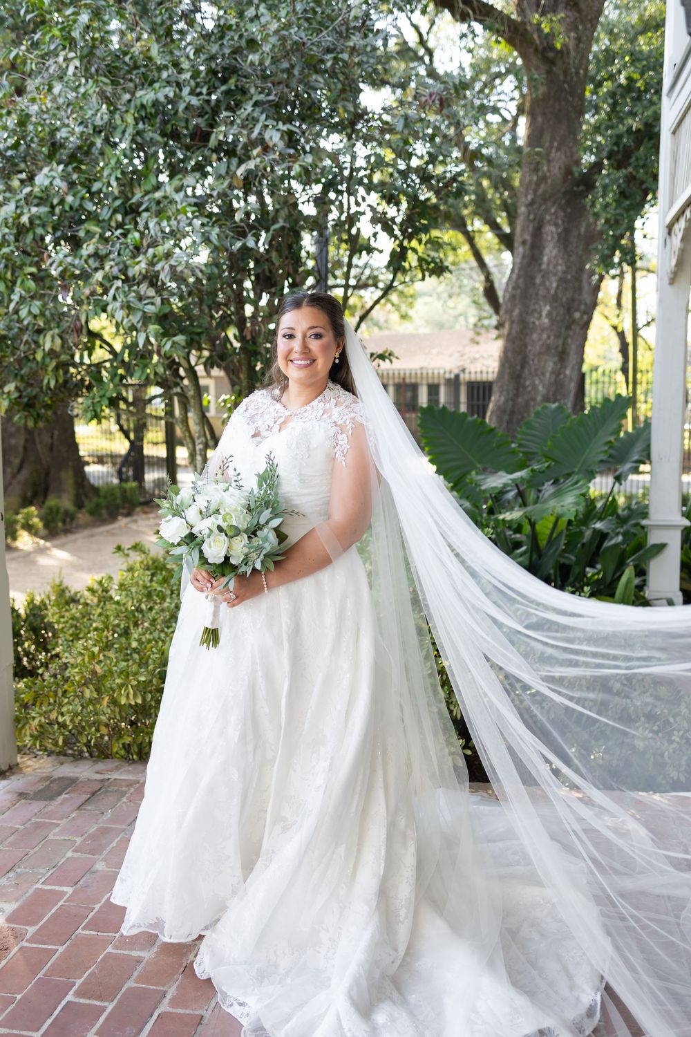 Bride in elegant white lace wedding dress with flowing cathedral veil holding white bouquet outdoors.