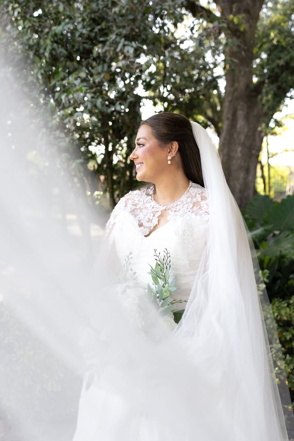 Smiling bride in lace long-sleeve wedding gown and veil holds bouquet outdoors among green trees.