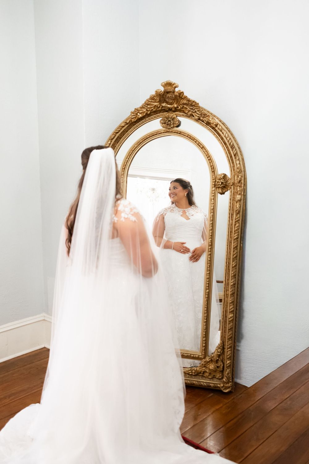 Bride in white lace gown and veil admiring her reflection in an ornate gold floor mirror.