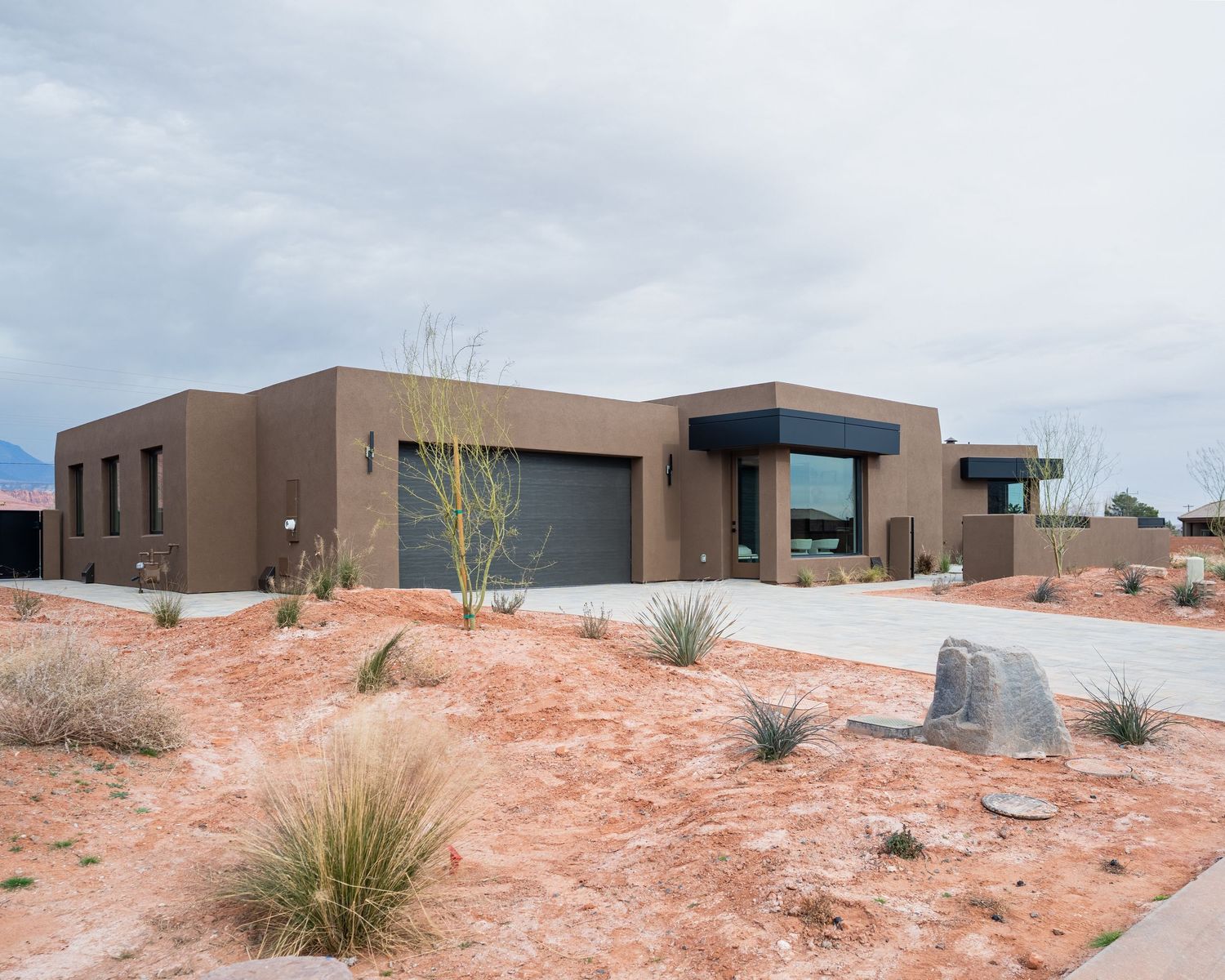 Modern desert home with flat roof, brown stucco exterior, and red sandy landscaping under cloudy skies.