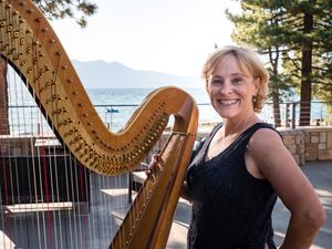 Smiling woman posing with a golden harp outdoors near a lake with mountains and trees in background.