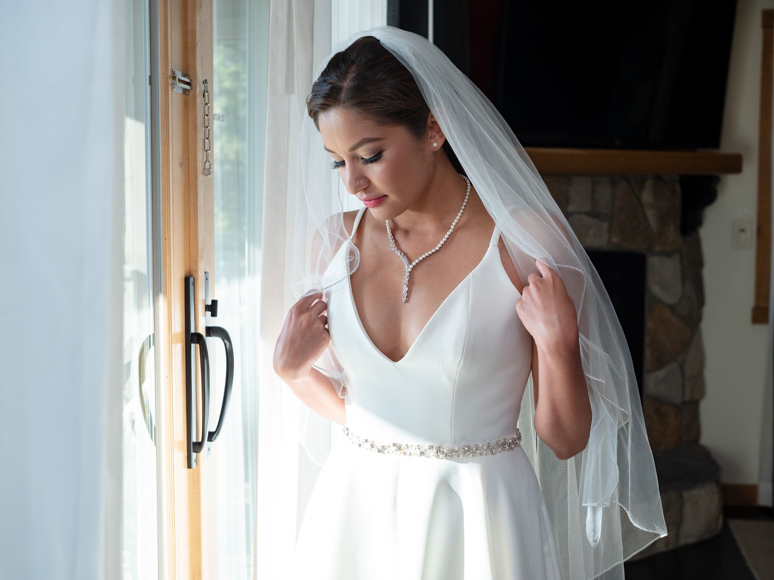 Bride in white wedding gown and veil standing by window, adjusting necklace on her wedding day.