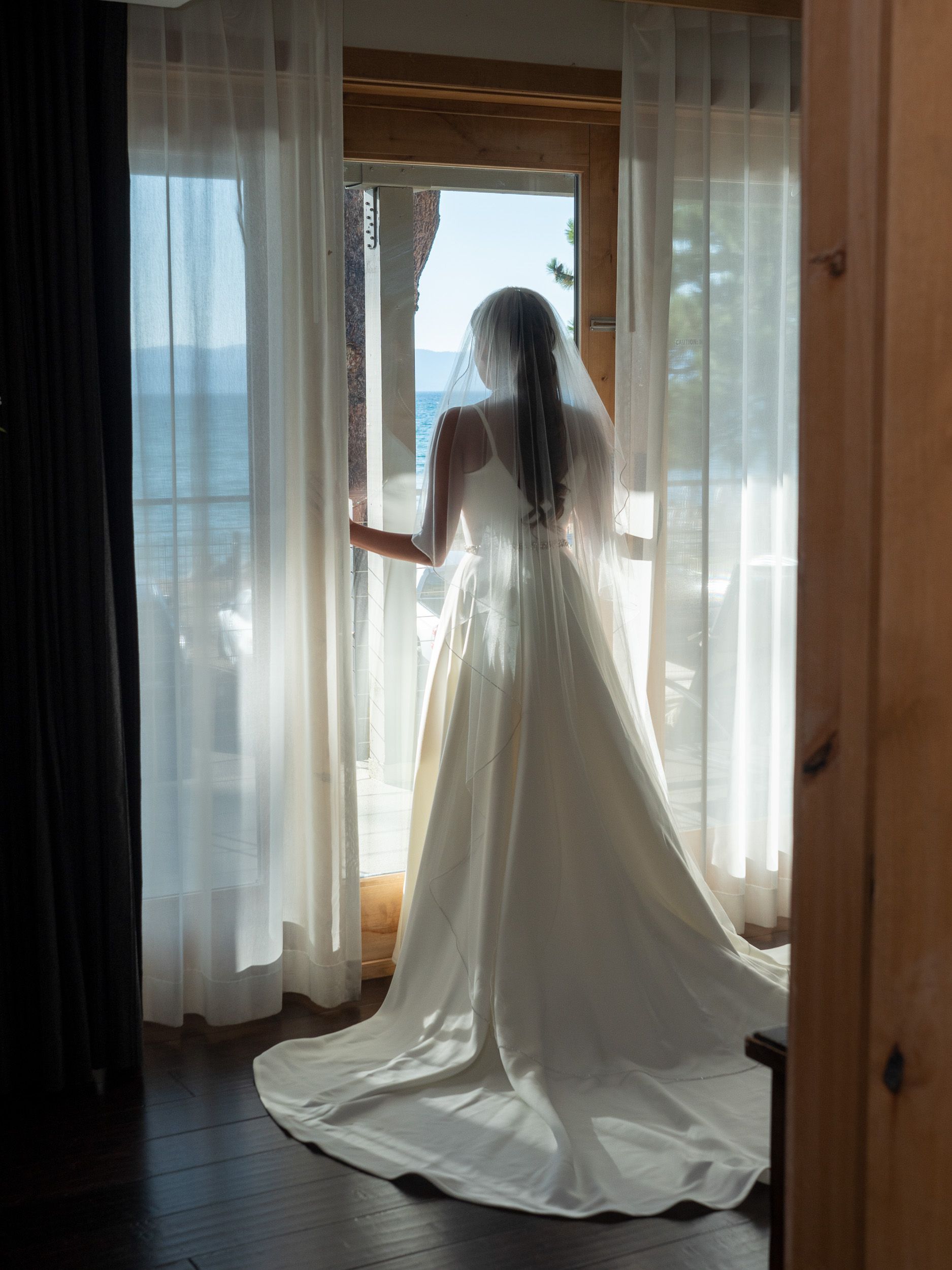 Bride in white wedding gown and veil stands at window overlooking ocean view.
