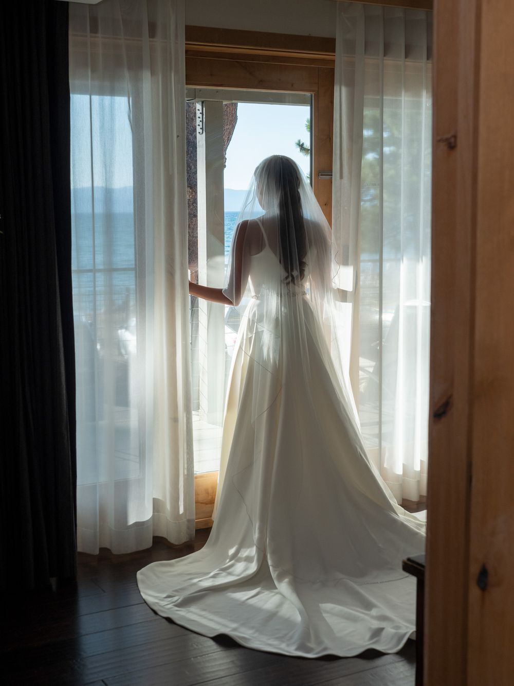 Bride in white wedding gown and veil stands at window overlooking ocean view.