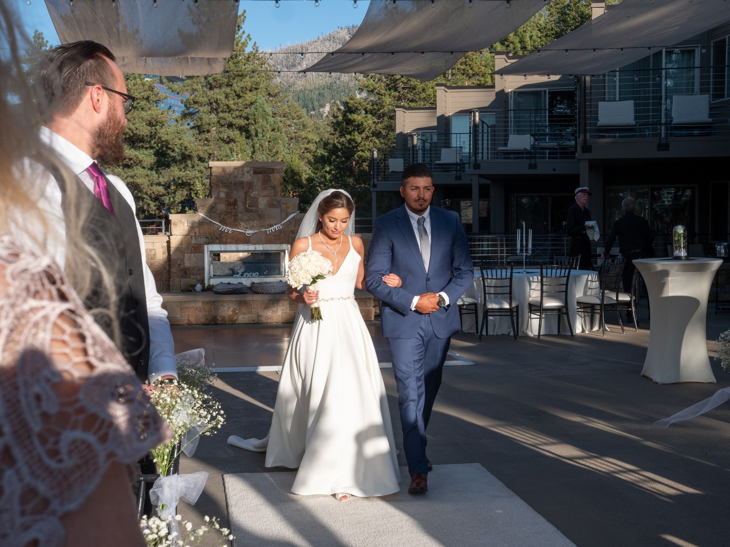 Bride and groom walking down outdoor wedding aisle at mountain venue, officiant waiting, guests gathered nearby.