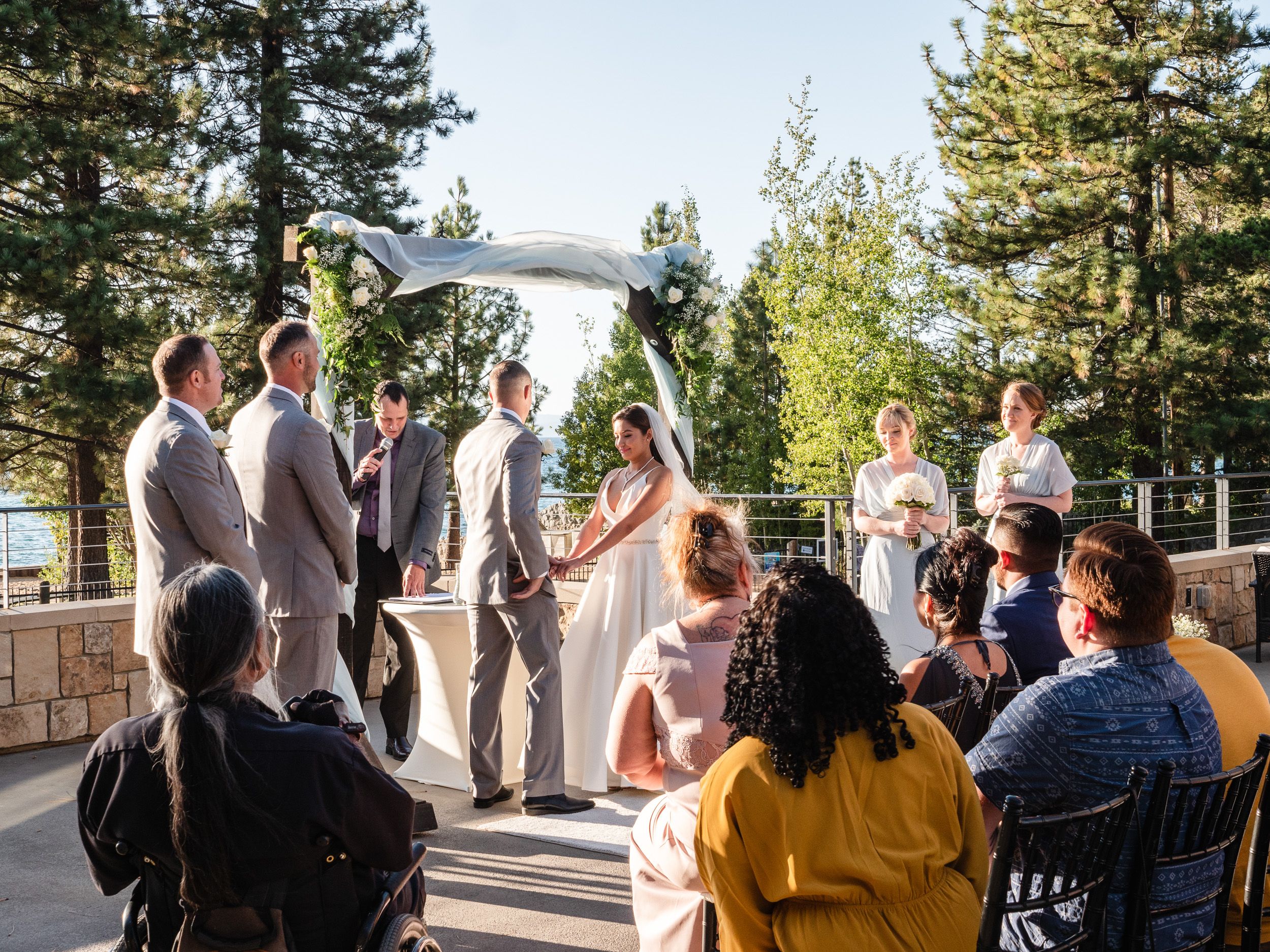 Outdoor wedding ceremony on a stone terrace surrounded by pine trees with a small gathering of guests watching the couple exchange vows.