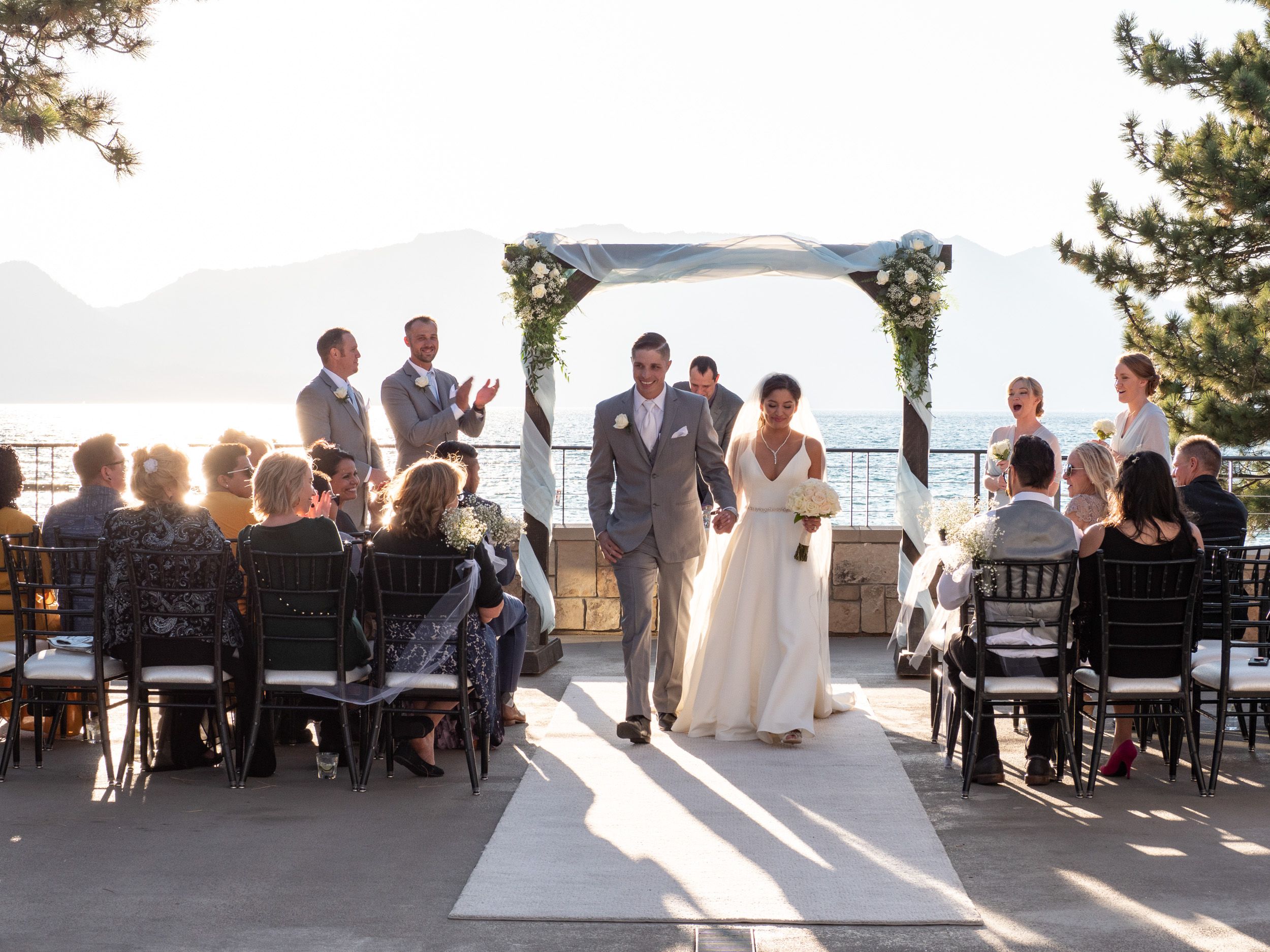 Bride and groom walk down the aisle at an outdoor lakeside wedding ceremony with seated guests.