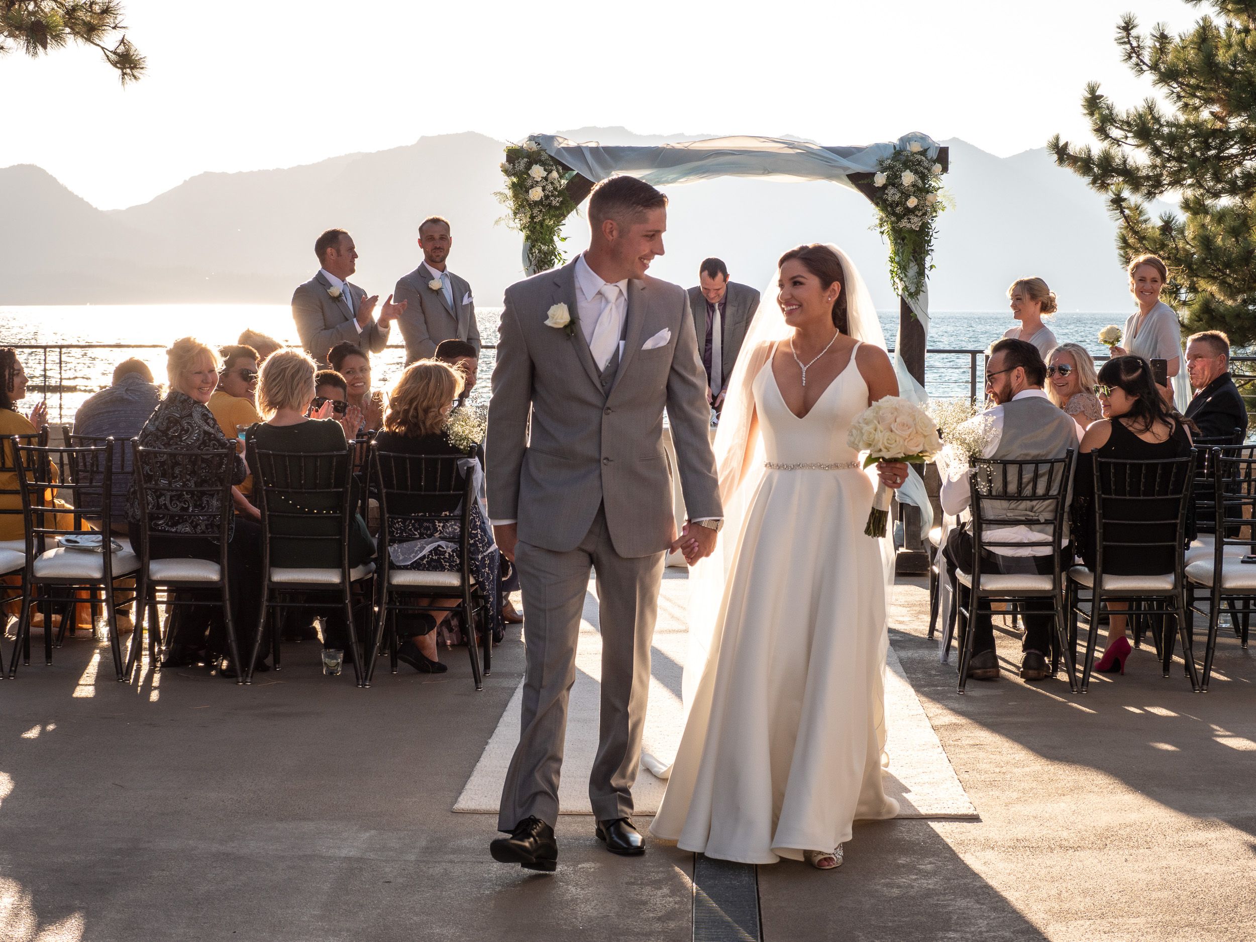 A bride and groom walk hand-in-hand after their outdoor lakeside wedding ceremony at sunset.
