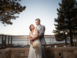 Bride and groom embrace on outdoor terrace with pine trees and lake at sunset.