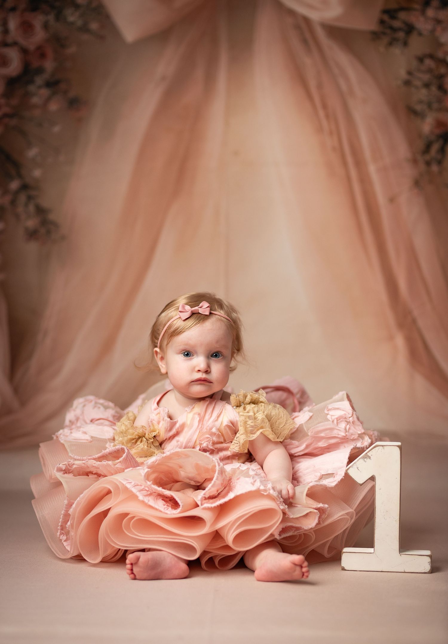 Baby girl in pink ruffle dress sits beside number 1 sign for first birthday portrait session.
