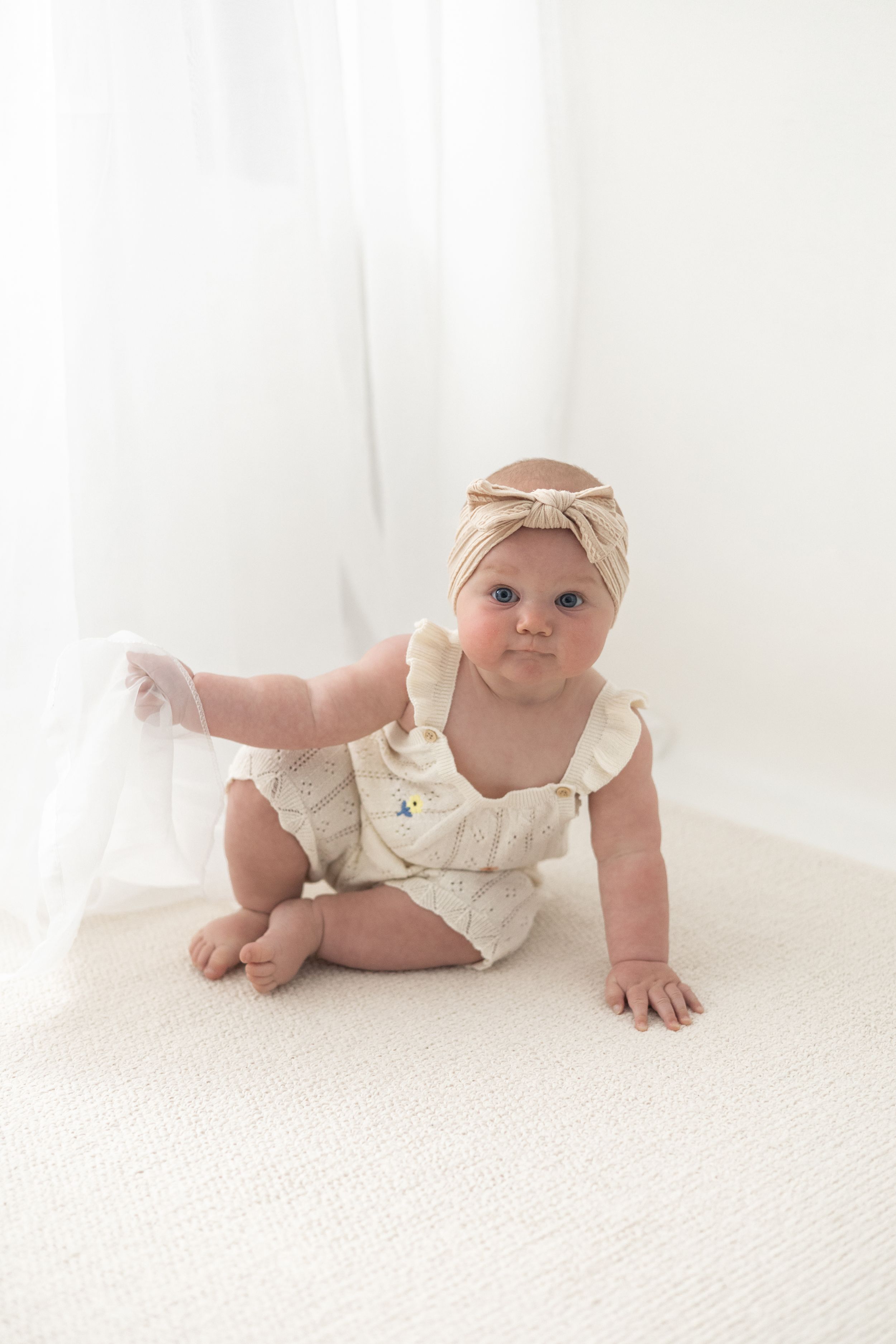 Baby girl in cream romper and tan bow headband crawling on white carpet in bright studio setting. Birthday photoshoot Nottingham
