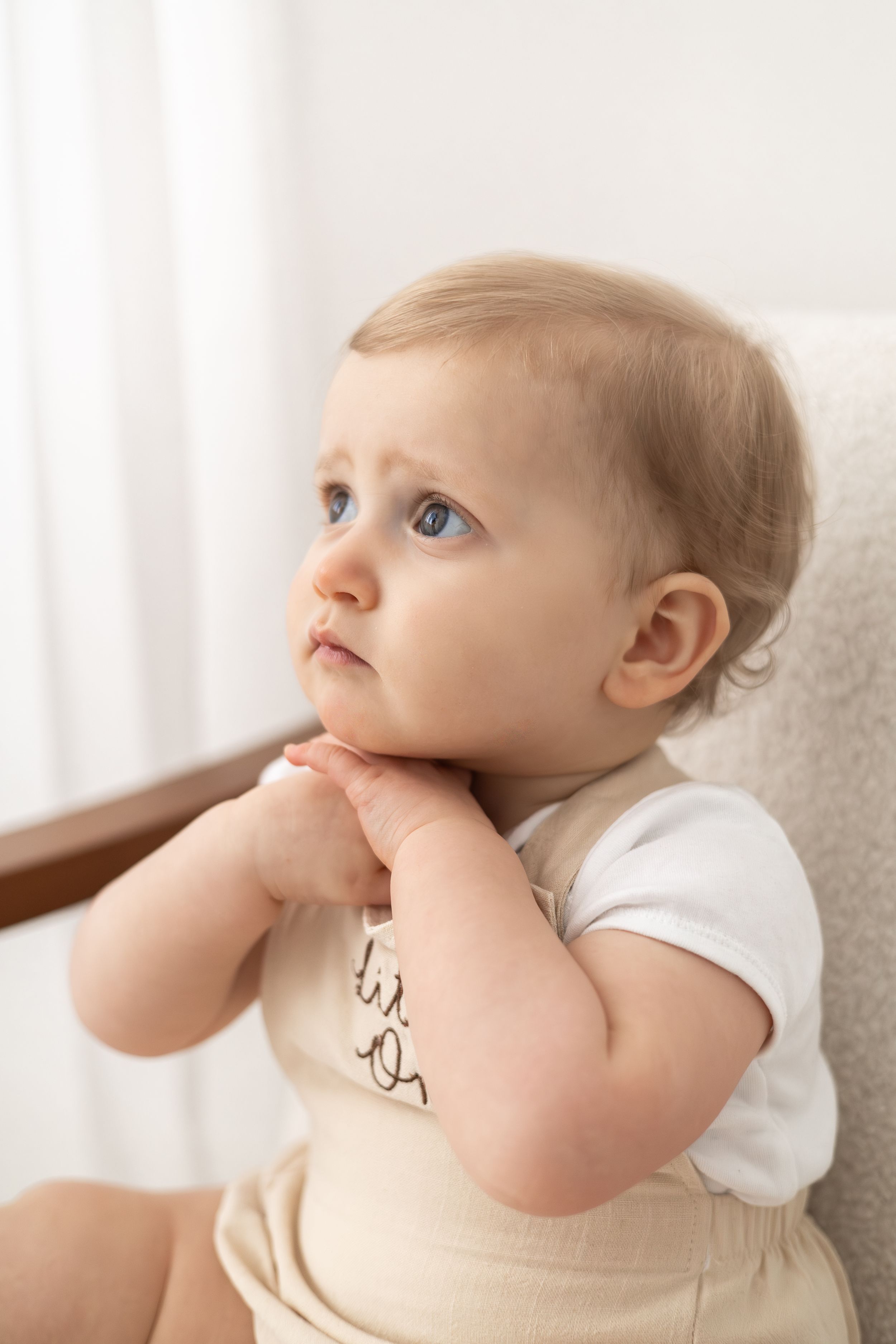 Adorable blue-eyed baby in white onesie and tan romper sitting in chair baby birthday photoshoot Nottingham