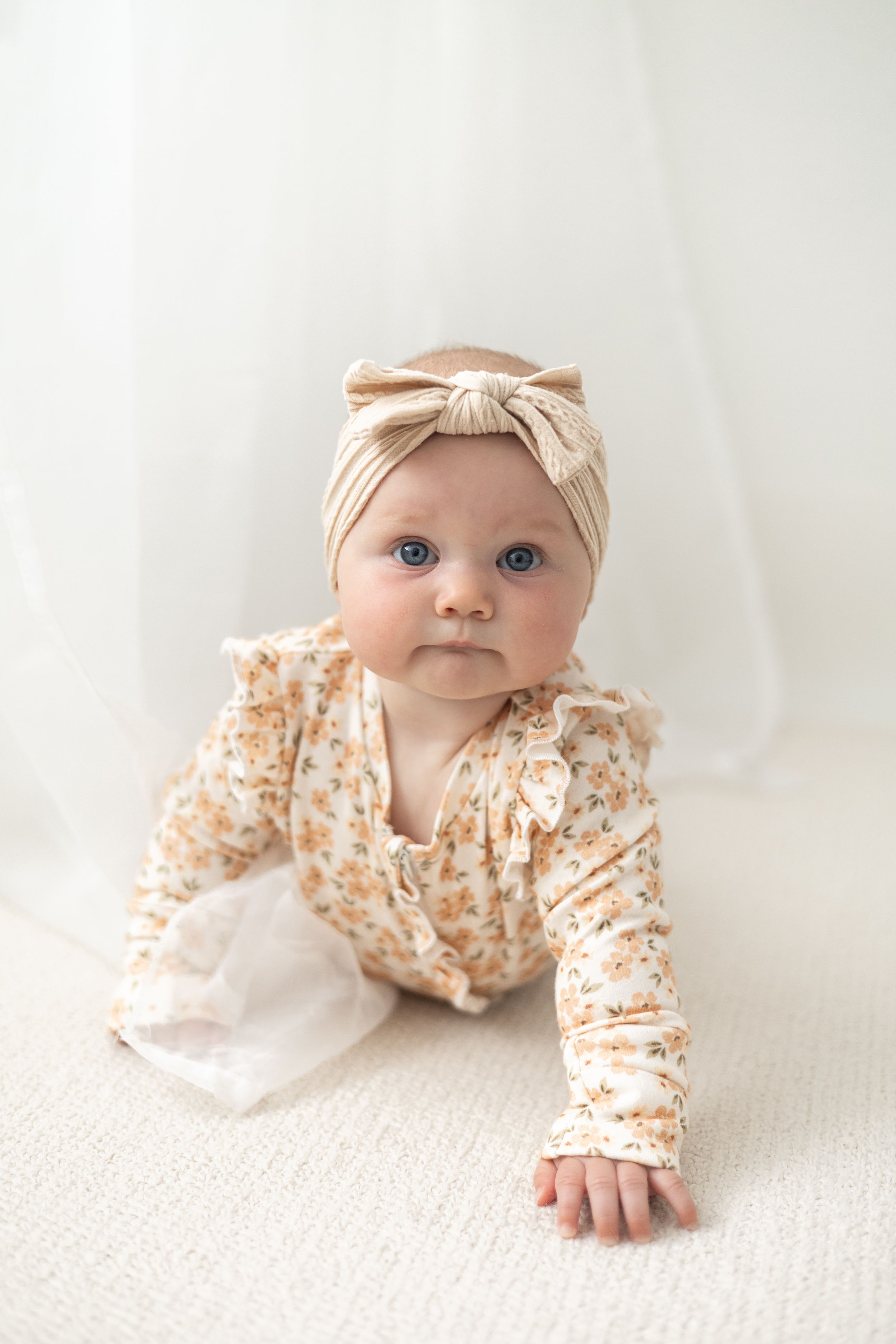 A baby girl in a floral romper and beige bow headband crawls on a white surface. Birthday photoshoot Nottingham