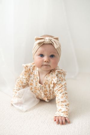 A baby girl in a floral romper and beige bow headband crawls on a white surface. Birthday photoshoot Nottingham