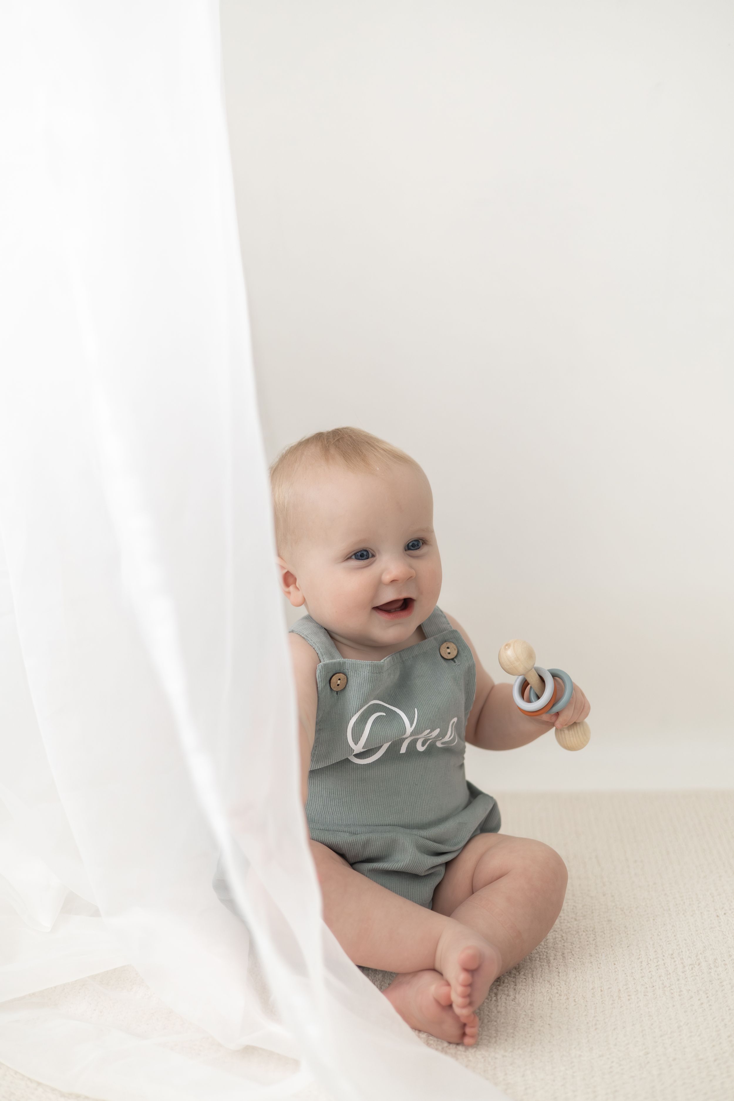 Smiling baby in sage green romper sits on white carpet holding a wooden toy rattle. Birthday photoshoot Nottingham