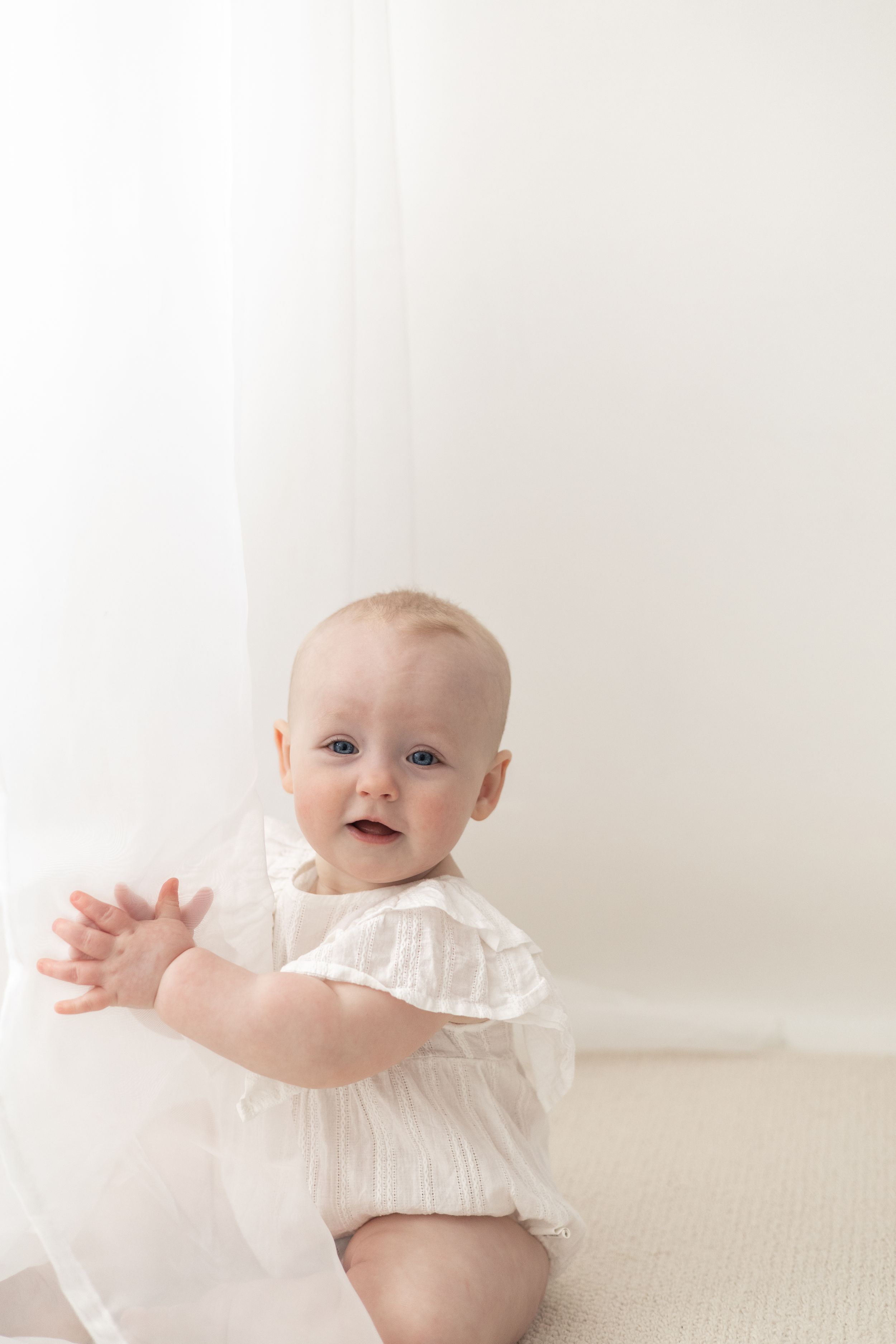 First birthday photography Nottingham, Smiling baby in white dress sitting on floor near sheer curtain in bright white room.