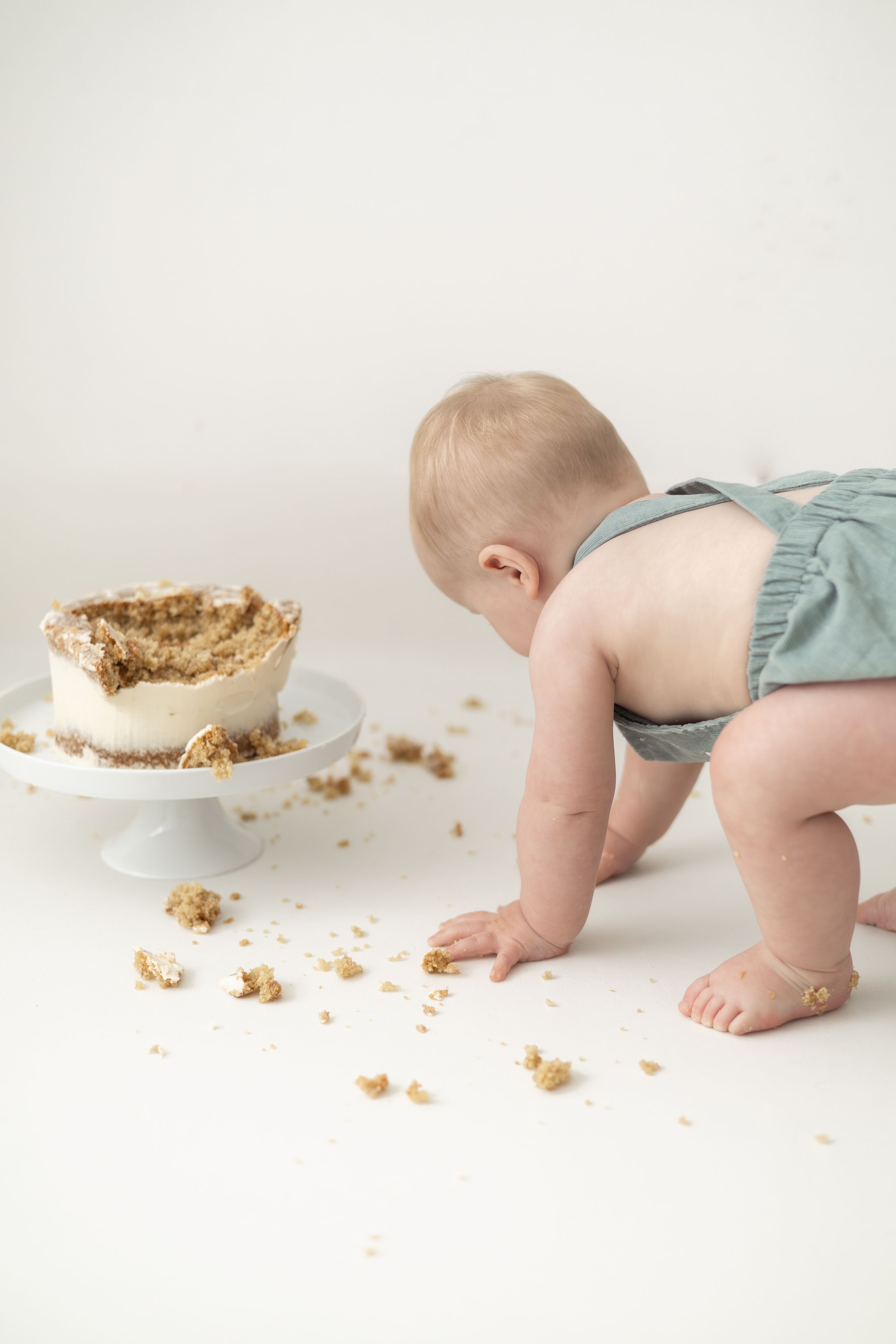 Baby in green romper smashing first birthday cake on white background, cake crumbles scattered on floor.  Birthday photoshoot Nottingham