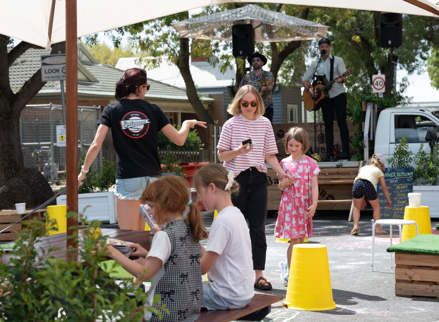 Children and adults enjoy an outdoor community event with live music, yellow buckets, and picnic seating on a sunny day.