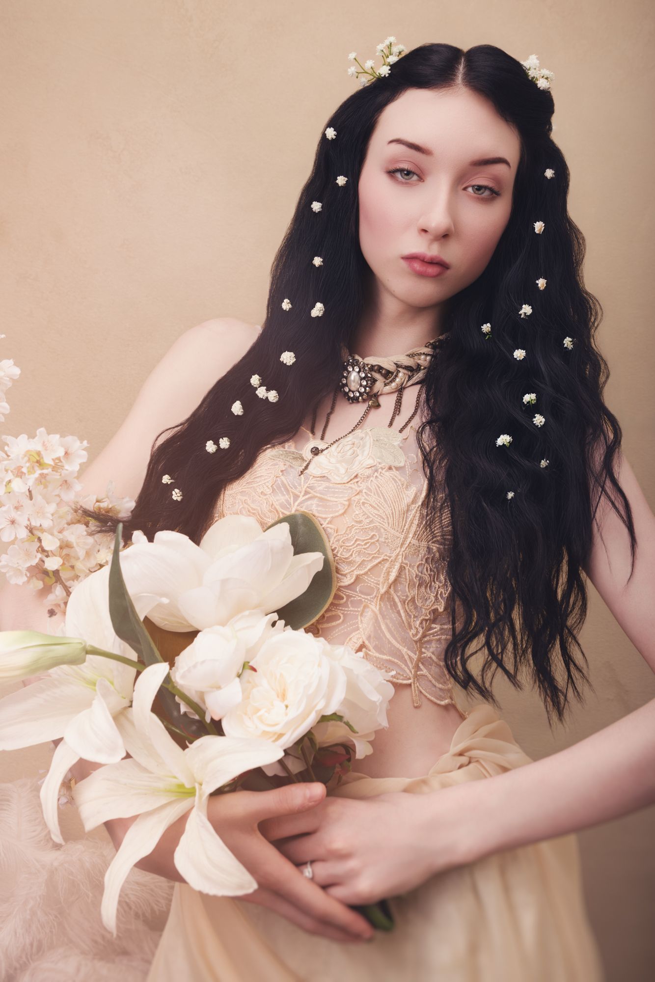 Young woman with long dark hair adorned with pearls, wearing a blush gown, holding white lilies and roses in a soft portrait.
