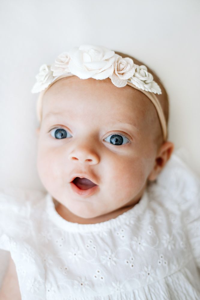 Adorable baby girl with blue eyes wearing a white dress and floral headband, looking up with a sweet expression.