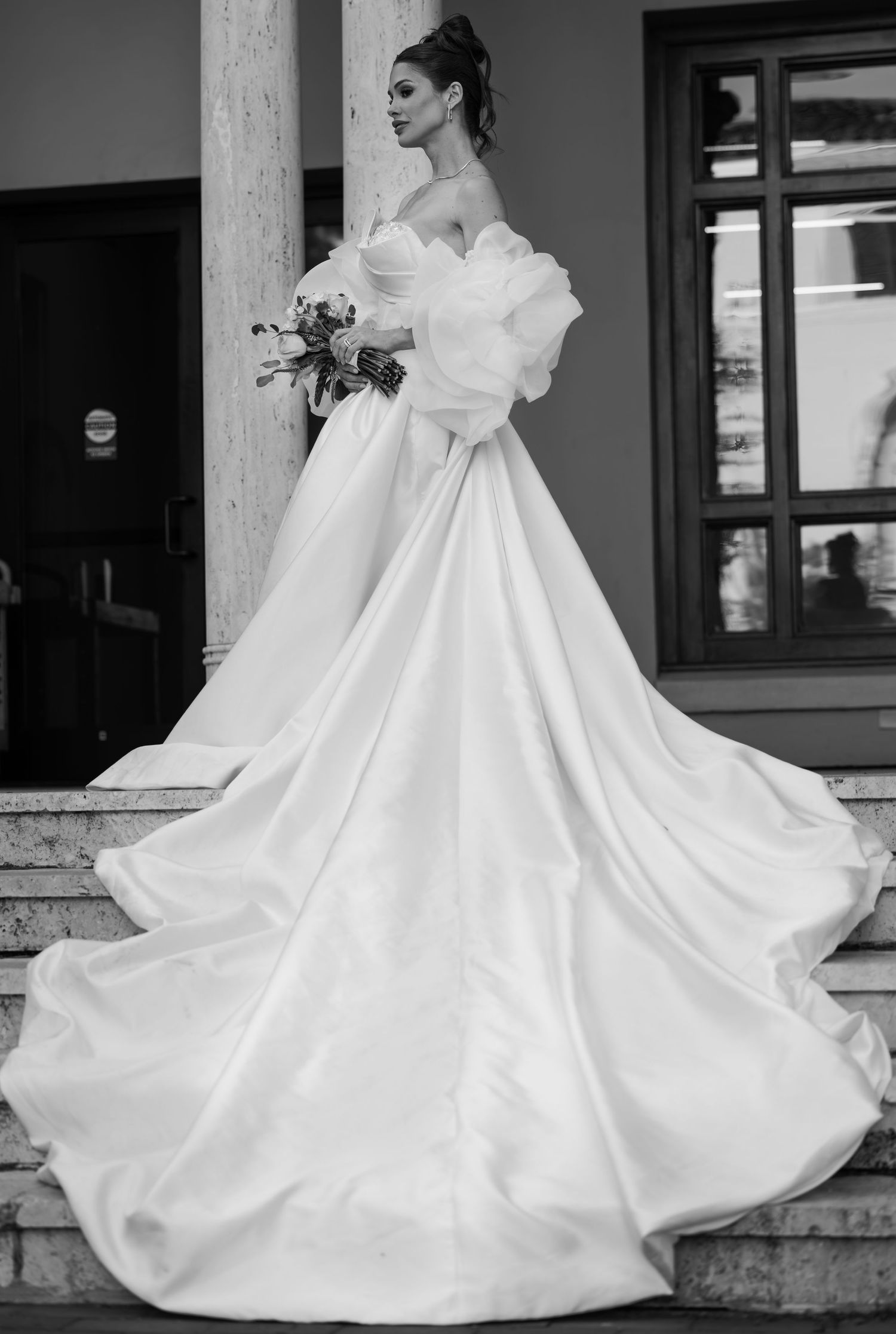 Bride in elegant off-shoulder ball gown with dramatic train and ruffle sleeves holds bouquet on stone steps.