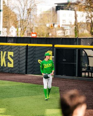 Oregon Ducks baseball player in green uniform standing in the outfield during a game at a college baseball stadium.