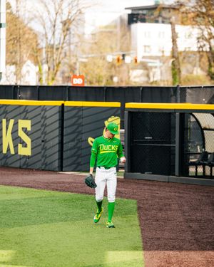 Oregon Ducks baseball player in green uniform standing in the outfield at a college baseball stadium.