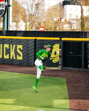 Oregon Ducks baseball player in green uniform throwing ball on outfield grass at PK Park stadium.