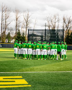 Oregon Ducks baseball team huddles together on a bright green field during a pre-game meeting in matching green uniforms.