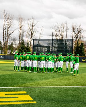 Oregon Ducks baseball team huddles together on the field in bright green uniforms during a cloudy day game.