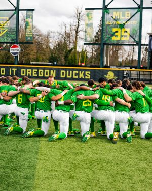 Oregon Ducks baseball team huddles together on the field in green uniforms, kneeling in a team circle before a game.