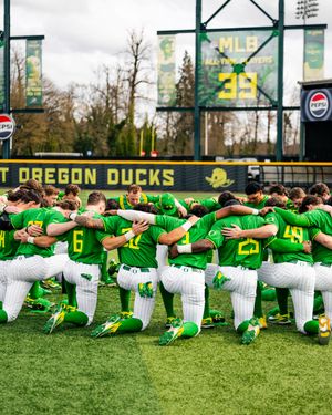Oregon Ducks baseball team kneeling together in a huddle on the field wearing green and white uniforms.