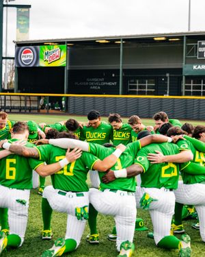Oregon Ducks baseball team huddles together in green uniforms on the field at their home stadium.