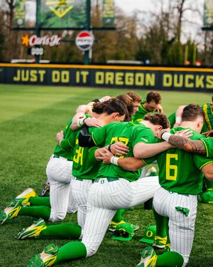 Oregon Ducks baseball team kneeling together in a huddle on the field, wearing green and white uniforms at PK Park.