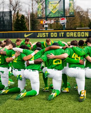 Oregon Ducks baseball team huddles together on the field in green uniforms at PK Park, kneeling in a team circle.