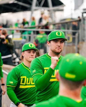 Oregon Ducks baseball player raises fist in dugout wearing green uniform and cap.