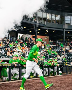 Oregon Ducks baseball player in green uniform walks onto field amid smoke during a game at a packed stadium.