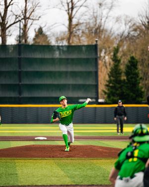 Oregon Ducks pitcher winds up to throw during a college baseball game at a green-fenced ballpark.