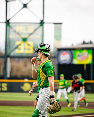 Oregon Ducks baseball catcher in green uniform with chest protector and mitt stands on field during a game.