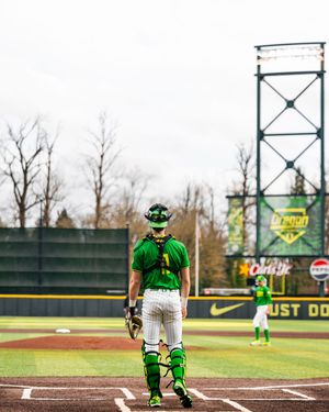 Oregon Ducks baseball catcher stands at home plate in green uniform and gear at PK Park stadium.