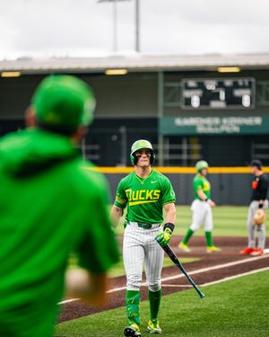 Oregon Ducks baseball player in green uniform holding bat walks toward teammate during a college baseball game.
