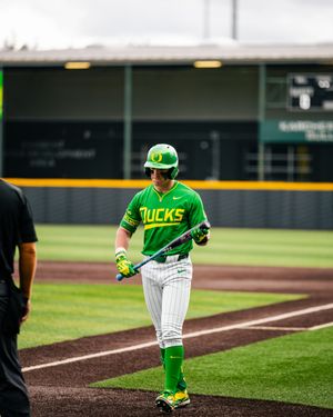 Oregon Ducks baseball player in green uniform walking on field holding bat during a college baseball game.